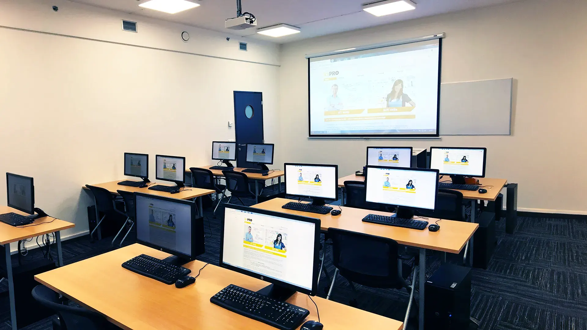 IT učebna se stoly, počítači s monitory a projekčním plátnem v přední části místnosti.