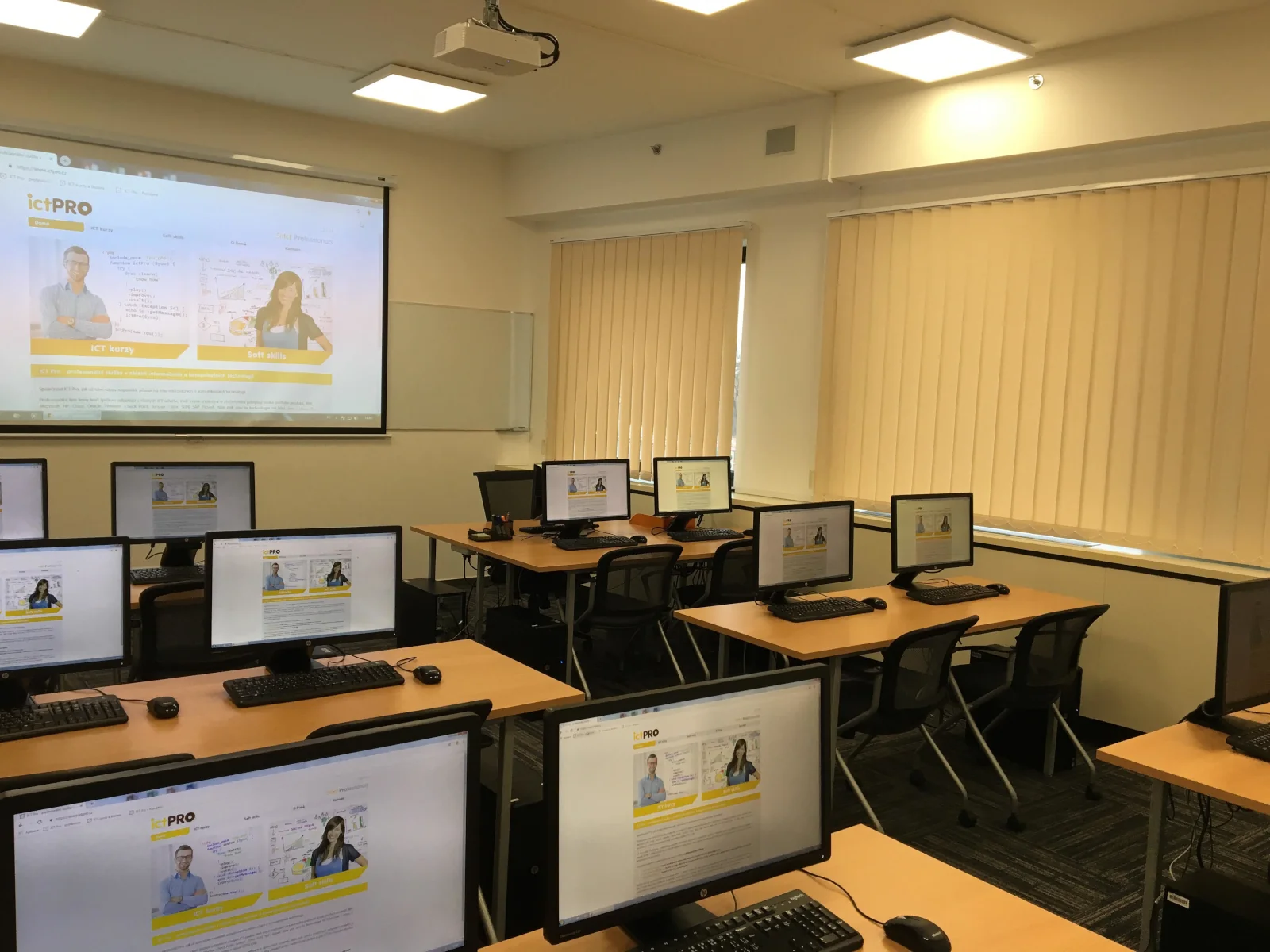Moderní počítačová učebna s monitory a projekčním plátnem připravená na IT školení.